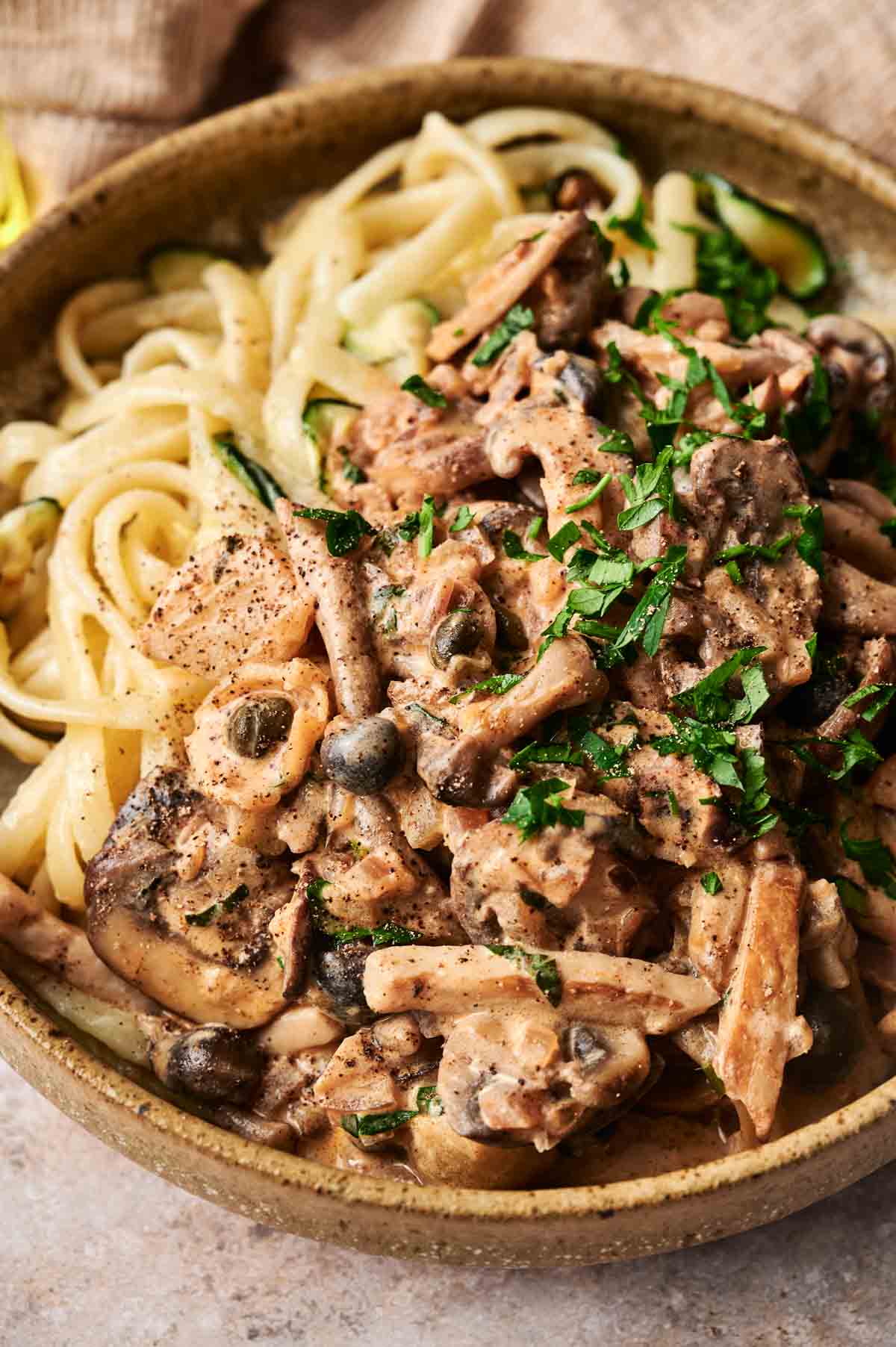 A bowl of pasta topped with a creamy mushroom stroganoff sauce, garnished with chopped herbs and black pepper.