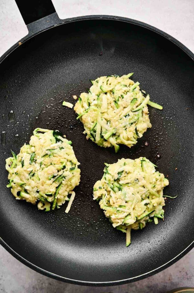 Three zucchini fritters sizzling in a black frying pan as they cook on the stovetop.