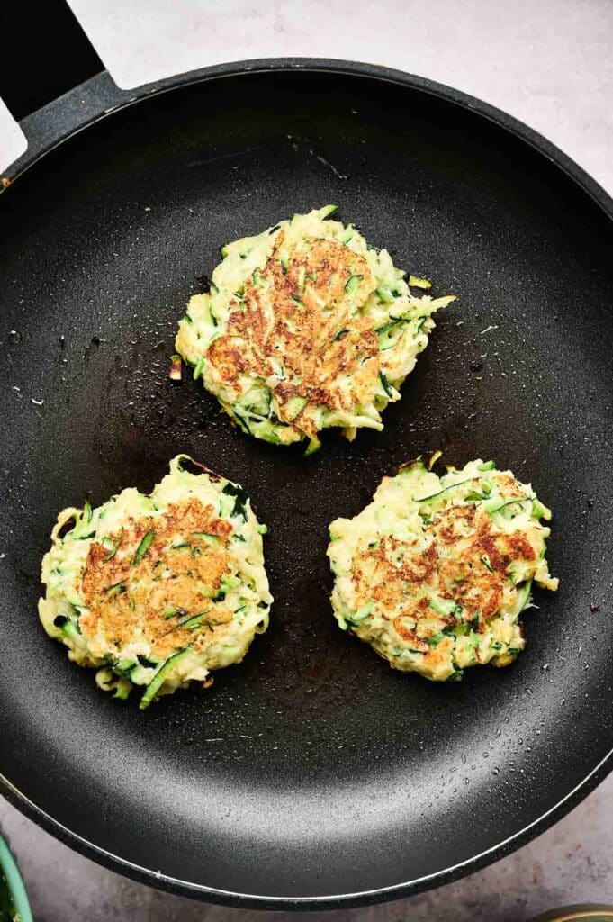 Three golden zucchini fritters sizzling in a black skillet, filling the air with their savory aroma.