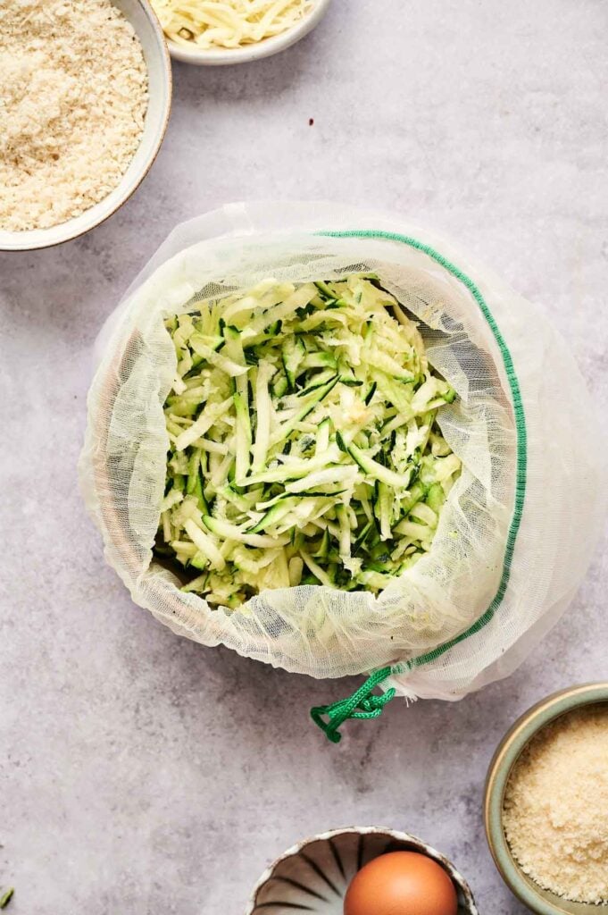 Shredded zucchini for fritters rests in a cloth over a bowl, surrounded by ingredients like breadcrumbs, cheese, and an egg on a light surface.