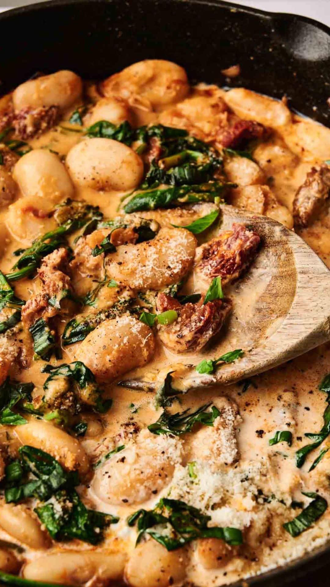 A creamy Marry Me Butter Beans dish shimmers in a pan, mingling sun-dried tomatoes and spinach, ready to be scooped with a wooden spoon.