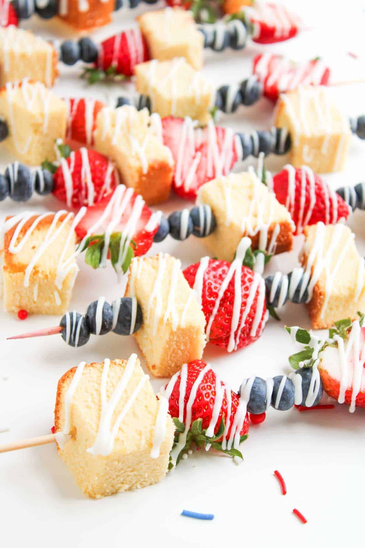 4th of July Fruit Kabobs: Wooden skewers with alternating pieces of pound cake, strawberries, and blueberries, drizzled with white icing and artfully arranged on a white surface.