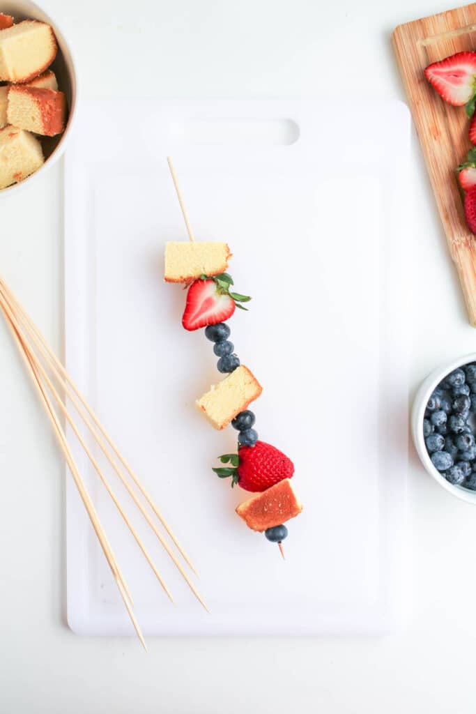 A wooden skewer threaded with cubes of pound cake, strawberries, and blueberries creates delightful 4th of July Fruit Kabobs, displayed on a white cutting board with extra ingredients nearby.