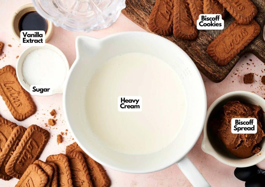 Mixing bowl of heavy cream surrounded by Biscoff cookies, Biscoff spread, sugar, and vanilla extract on a pink surface&mdash;perfect ingredients for making creamy Biscoff Ice Cream.