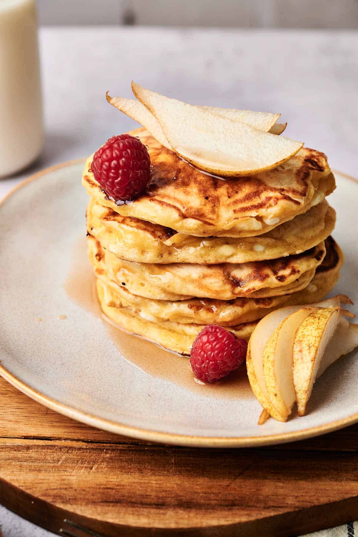 A stack of cottage cheese pancakes topped with pear slices and raspberries is beautifully presented on a plate, drizzled with syrup. A glass of milk stands invitingly in the background.