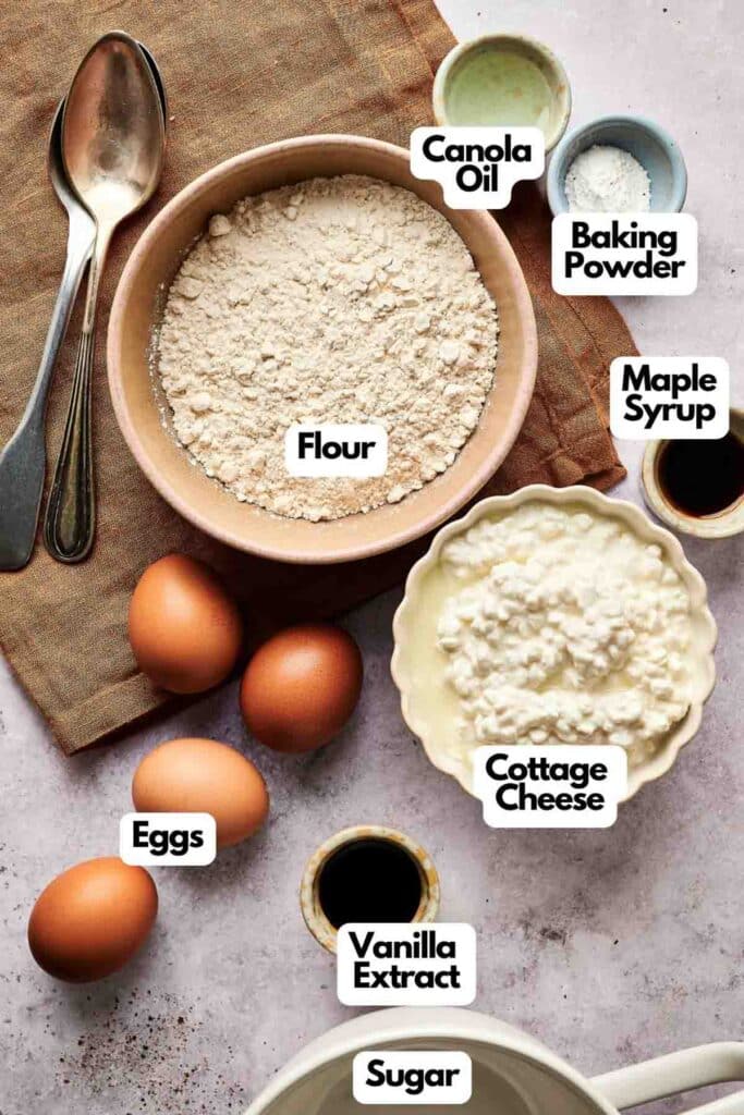 Baking ingredients for cottage cheese pancakes are artfully arranged on a table, featuring flour, canola oil, baking powder, maple syrup, eggs, vanilla extract, and sugar.
