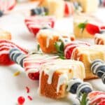4th of July Fruit Kabobs with cubes of pound cake, strawberries, and blueberries, drizzled with white icing, arranged on a white surface.