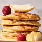 A stack of cottage cheese pancakes topped with pear slices and a raspberry, with additional pear slices and raspberries nestled at the base.