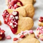 Heart-shaped cookies with pink and red icing are beautifully adorned with heart and star sprinkles, elegantly displayed on a white surface.