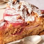 A slice of rhubarb cake dusted with powdered sugar sits on a plate, accompanied by a silver spoon.