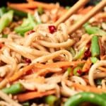 Close-up of chopsticks holding spicy noodles stir-fried with sliced carrots, green beans, red chili, and sesame seeds.