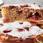 A slice of strawberry cake is held up above the rest, its powdered sugar glistening. Plates are stacked in the background, ready for more servings.