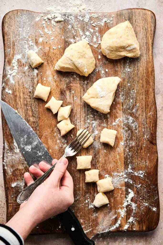 Person shaping gnocchi dough into pieces on a floured wooden board with a knife and fork.