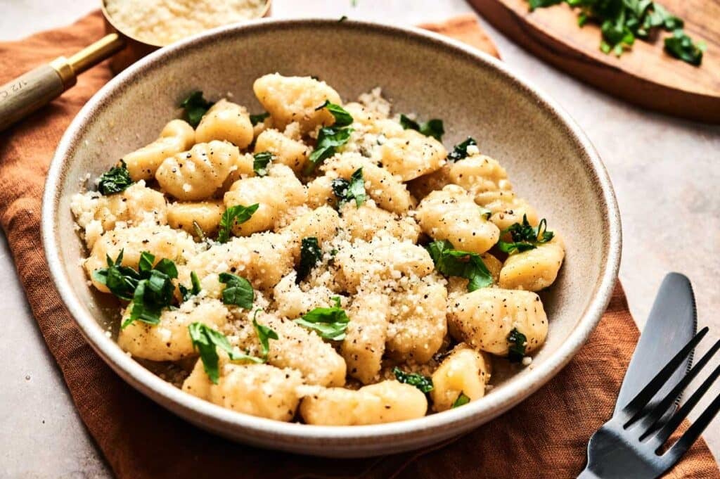 A delightful bowl of gnocchi crowned with grated cheese, a hint of black pepper, and fresh parsley rests on a brown napkin. Adjacent lie a fork and spoon, while in the background await additional bowls of cheese and parsley for those who crave an extra dash of flavor.