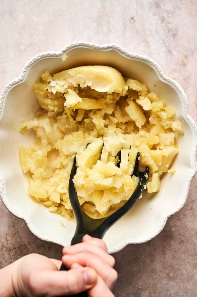 Hands expertly mashing boiled potatoes in a white bowl with a black masher on a beige surface, preparing the perfect base for homemade gnocchi.