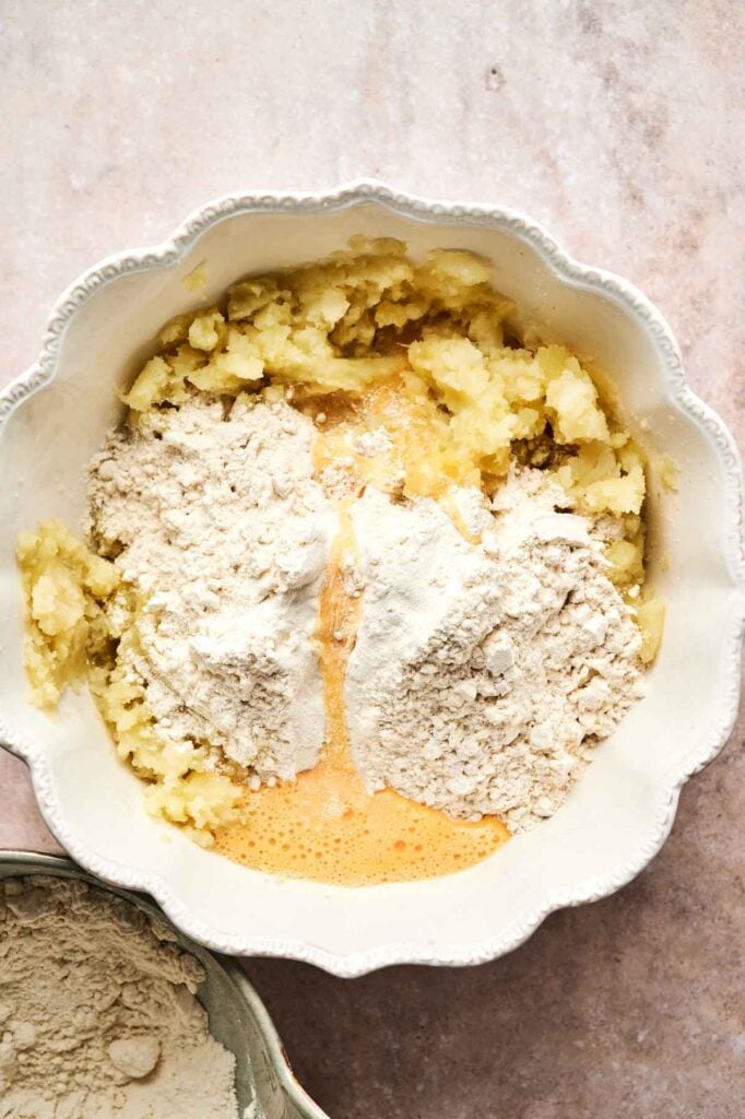 A mixing bowl containing mashed potatoes, flour, and a whisked egg sits on a marble surface, ready to transform into delicious homemade gnocchi.