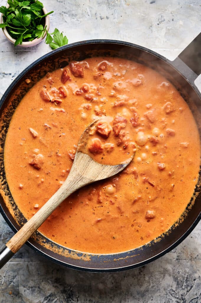 A wooden spoon rests in a frying pan filled with creamy tomato sauce, garnished with chopped herbs on top as it simmers into a delightful Halloumi Stroganoff.