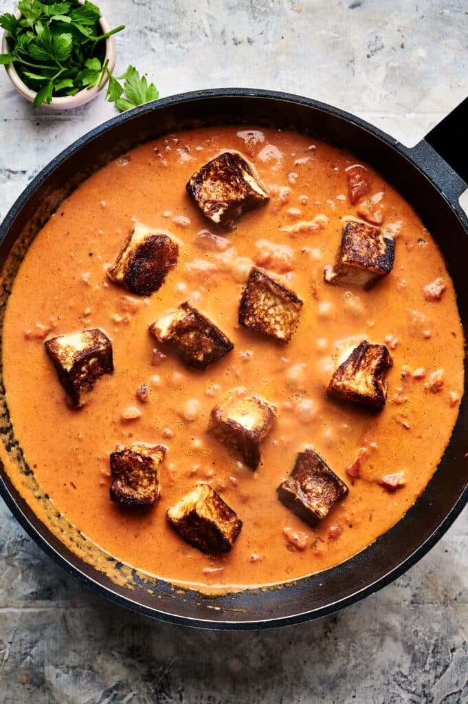 Black skillet with a creamy tomato curry sauce and grilled paneer cubes, reminiscent of a Halloumi Stroganoff. Fresh herbs in a small bowl nearby.