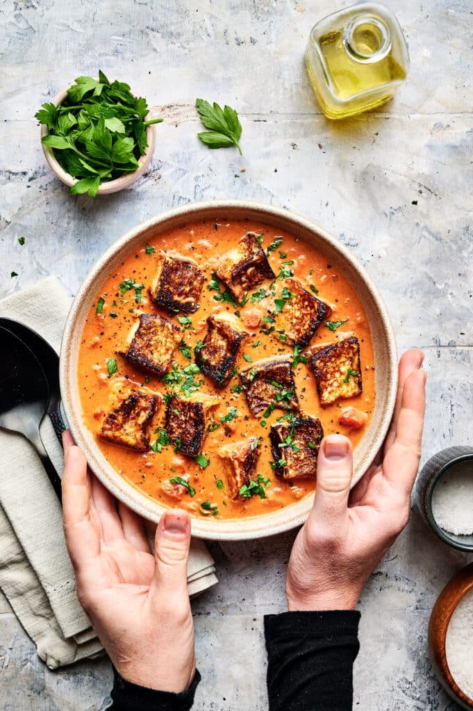 Hands holding a bowl of tomato soup topped with toasted croutons and herbs, next to a small bottle of olive oil and fresh parsley on a textured surface, evoke the comfort of halloumi stroganoff with every aromatic swirl.