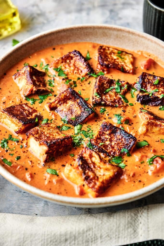 A bowl of creamy tomato curry with pieces of seared paneer, reminiscent of a Halloumi Stroganoff, garnished with chopped herbs and displayed on a textured surface.