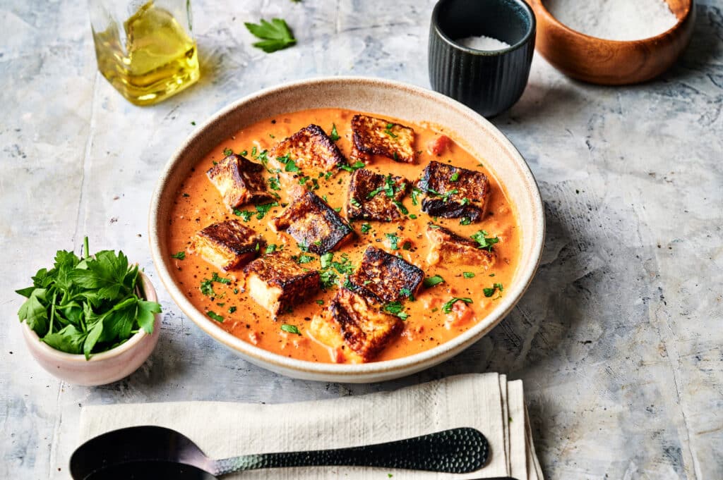 A bowl of tomato soup with grilled cheese croutons, garnished with herbs, sits ready to enjoy. Halloumi Stroganoff-inspired seasonings add a unique twist. Spoons and a napkin are in the foreground; oil and more flavors await in the background.