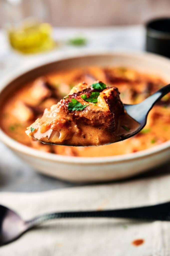 Close-up of a spoon holding a piece of bread covered in creamy, orange Halloumi Stroganoff, garnished with herbs. A bowl filled with the same savory delight is in the background.