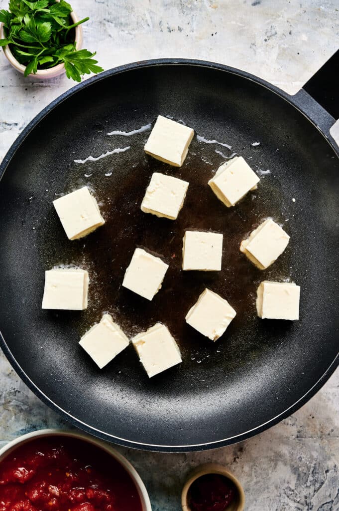 Cubes of tofu frying in a non-stick pan with oil, reminiscent of halloumi stroganoff. A small bowl of tomato sauce and a cup of parsley are nearby.