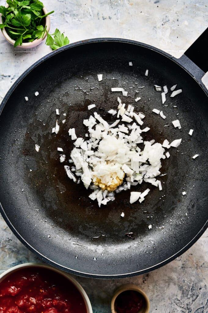 Chopped onions and garlic are being sautéed in a black pan, surrounded by ingredients in small bowls, including tomatoes and herbs—creating the perfect base for a delectable Halloumi Stroganoff.