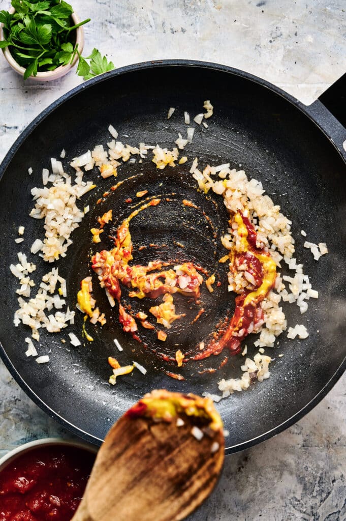 Pan with sautéed onions, tomato paste, and a spoon for stirring the creamy Halloumi Stroganoff.