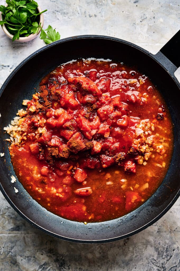 A skillet filled with tomato sauce, chopped tomatoes, and grains sits on a textured surface, reminiscent of a hearty Halloumi Stroganoff. A small bowl of parsley adds a fresh touch nearby.