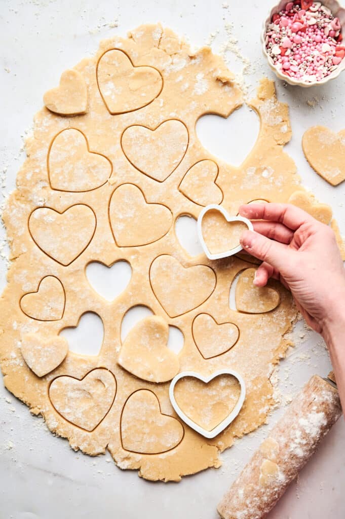 A hand uses a heart-shaped cookie cutter on rolled-out dough, creating perfect heart-shaped cookies. Nearby, a bowl of pink and white sprinkles waits to add a whimsical touch.