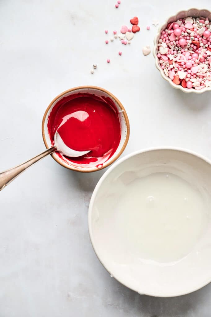 Two bowls with icing, one red and one white, sit on a white surface beside heart-shaped cookies. A spoon rests in the vibrant red icing, ready for spreading. Nearby, a bowl of colorful sprinkles awaits its turn to add an extra touch of sweetness to the scene.