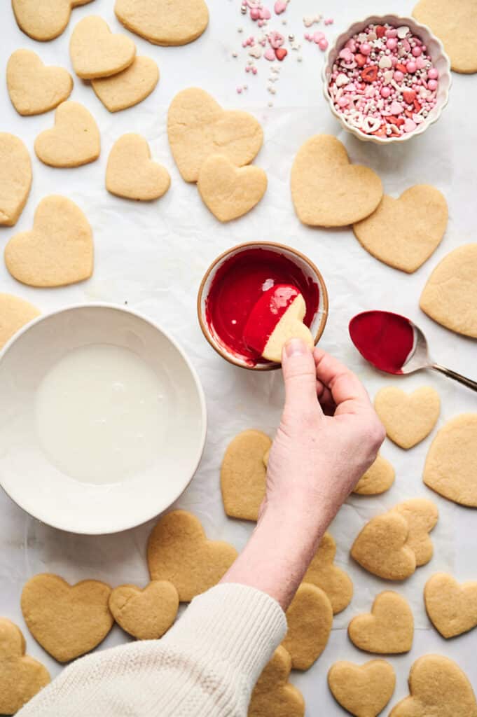 A person dips heart-shaped cookies into red icing. Surrounding the bowl are other plain cookies, a bowl of white icing, and a bowl of decorative sprinkles. The process brings an artistic flair to each treat, transforming them into delightful edible creations bursting with love.
