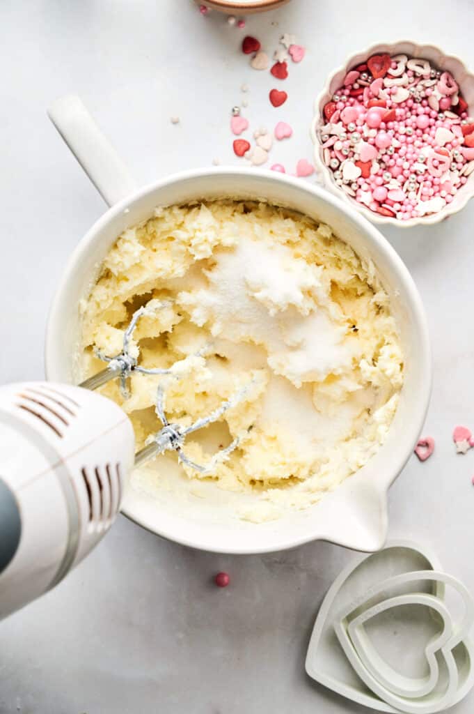A mixing bowl brimming with batter and sugar, surrounded by heart-shaped sprinkles and cookie cutters, while an electric hand mixer whirs away, setting the stage for delightful heart-shaped cookies.