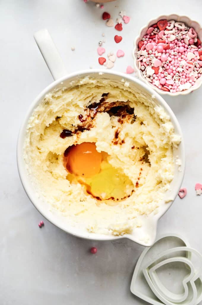 A mixing bowl with creamed butter and sugar, vanilla, and two eggs awaits transformation into heart-shaped cookies. Nearby, a cookie cutter and heart-shaped sprinkles rest on the marble surface, ready to add that extra touch of love.