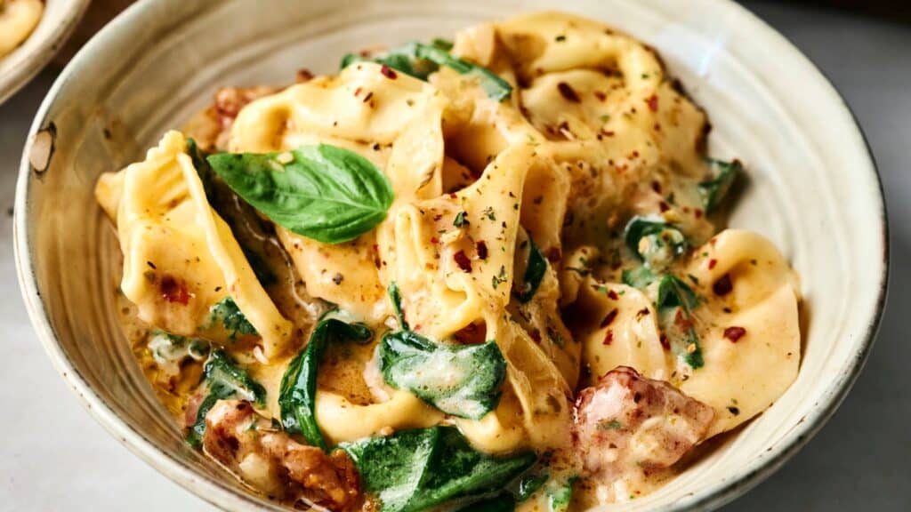 A bowl of creamy tortellini pasta with spinach, sun-dried tomatoes, herbs, and a basil garnish.