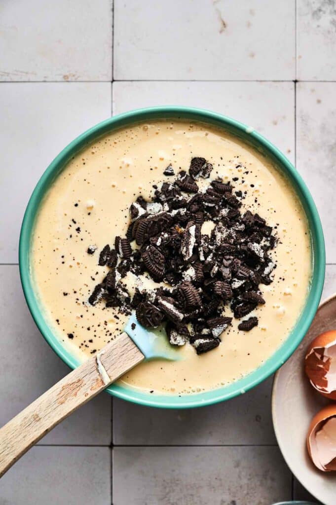 A bowl of creamy yellow batter is topped with crushed chocolate cookies, reminiscent of an Oreo cheesecake, accompanied by a spatula. It sits elegantly on a tiled surface, surrounded by scattered eggshells.