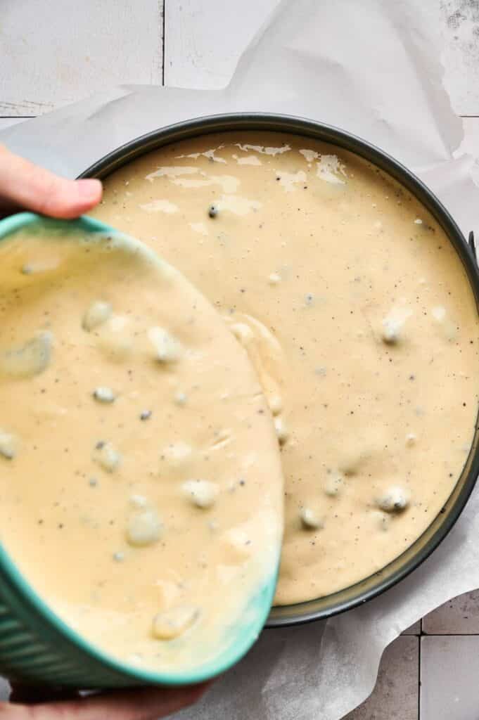Pouring creamy Oreo cheesecake batter with visible chunks into a round baking pan lined with parchment paper.