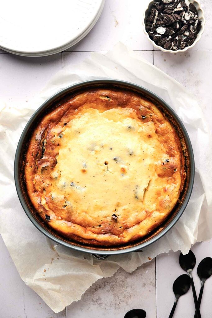 An Oreo cheesecake with chocolate chips sits on parchment paper in a round pan. A bowl of crushed cookies and three spoons are nearby, ready to dive into the creamy delight.