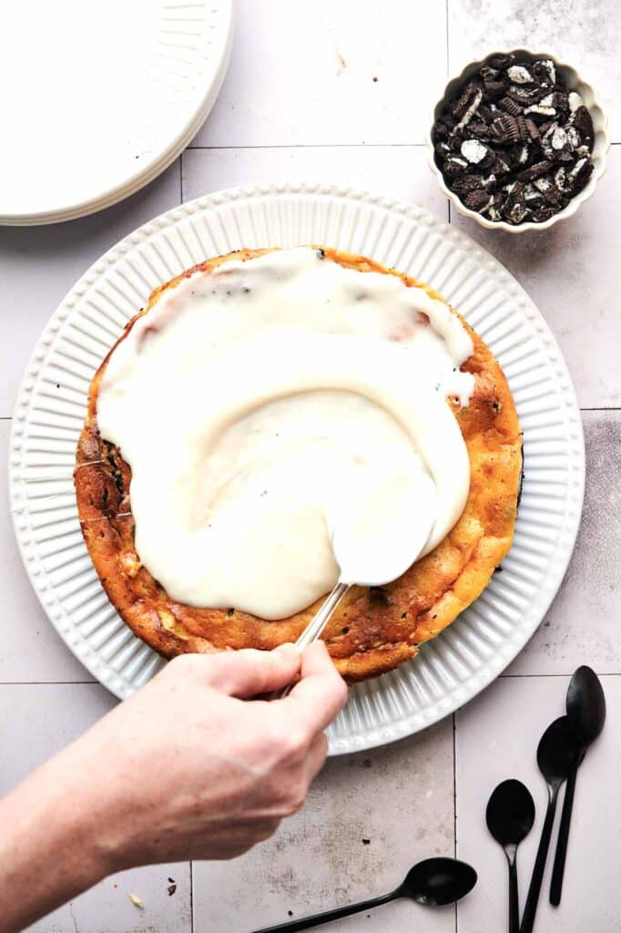 A hand spreads white frosting over a round cake on a white plate, crafting the perfect Oreo cheesecake delight. Three black spoons sit nearby, alongside a small bowl of crumbled cookies in the background.