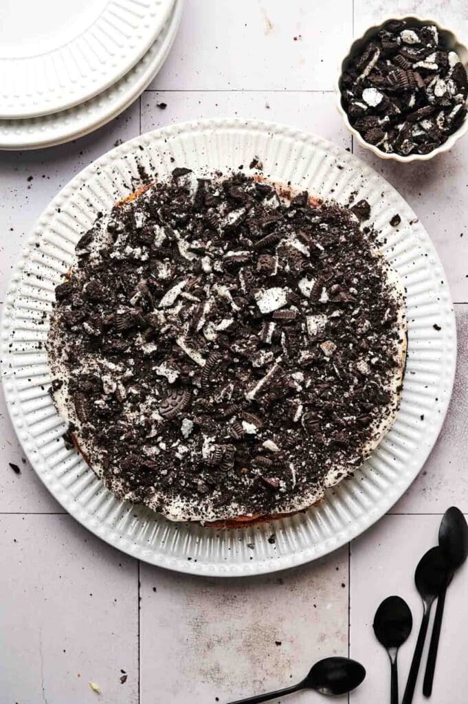 Oreo cheesecake topped with crushed cookies sits elegantly on a white plate, accompanied by a bowl of extra crushed cookies and a few black spoons nearby.