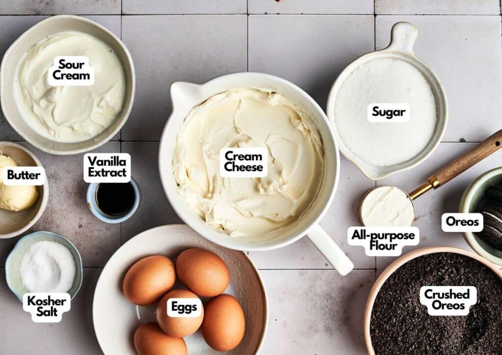 A flat lay of labeled cheesecake ingredients including sour cream, vanilla extract, butter, kosher salt, eggs, cream cheese, sugar, all-purpose flour, Oreos, crushed Oreos, and a hint of Biscoff Ice Cream inspiration.