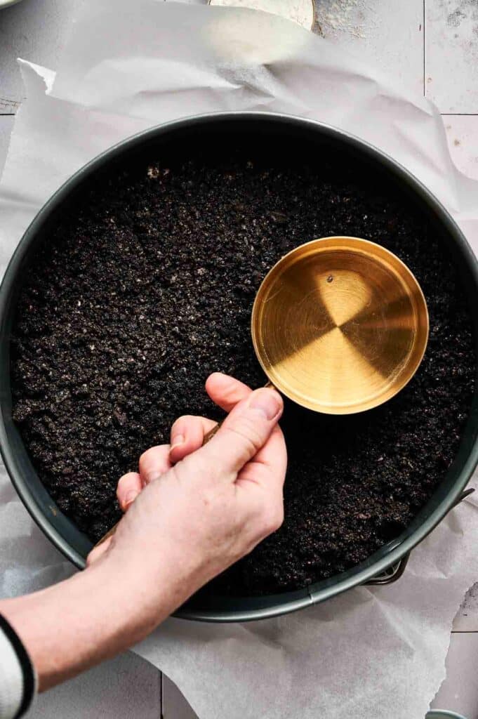 A hand uses a brass measuring cup to flatten a layer of crushed cookie crust, perfect as the base for an indulgent Oreo cheesecake, in a round baking pan lined with parchment paper.