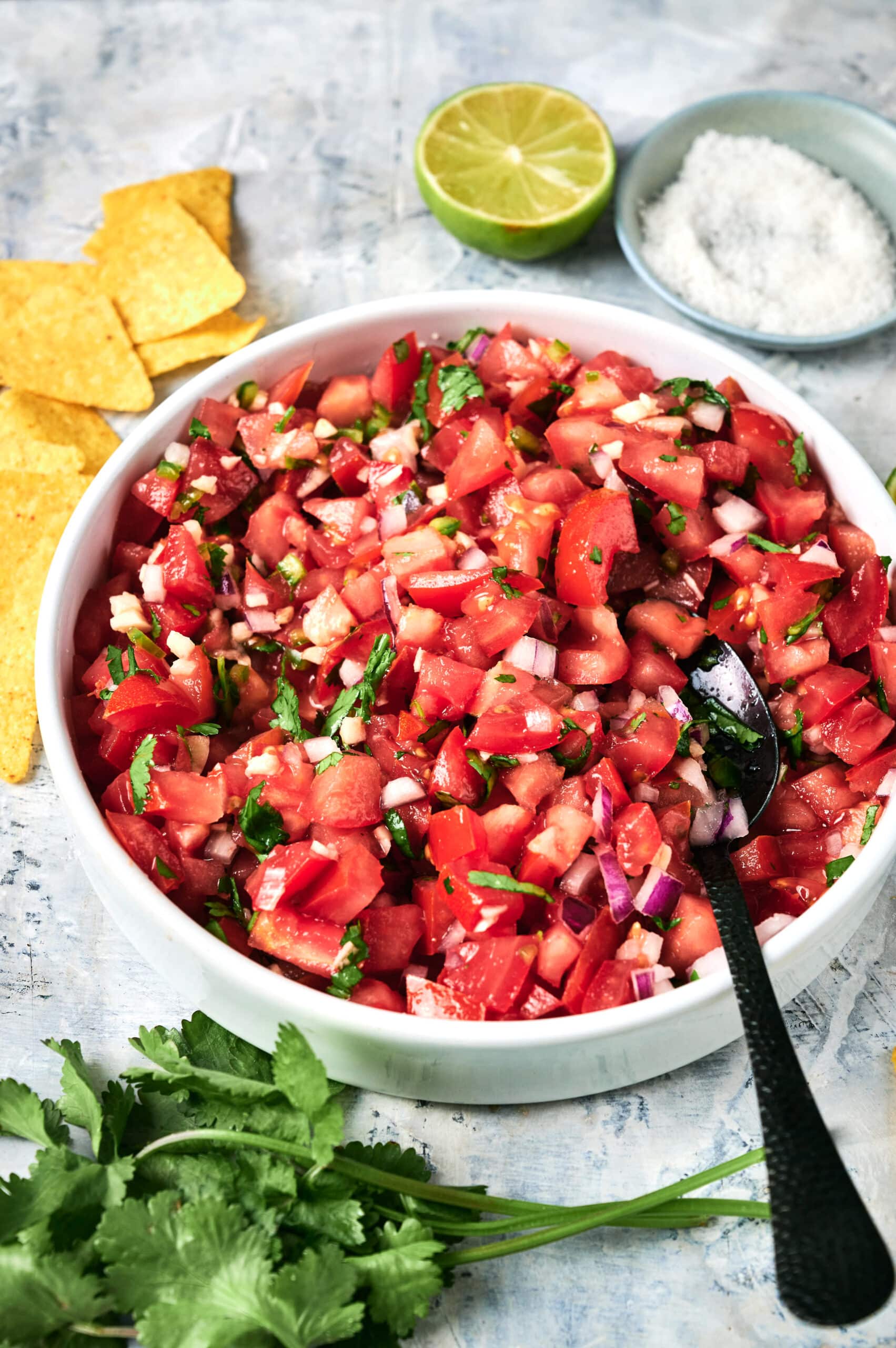 A bowl of freshly made Pico de Gallo with diced tomatoes, onions, cilantro, and jalape&ntilde;o sits surrounded by tortilla chips, lime wedges, salt, and fresh cilantro.