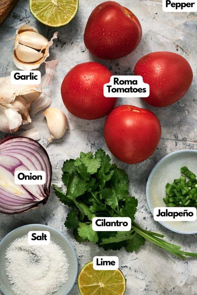 Ingredients for pico de gallo are arranged on a surface, including roma tomatoes, garlic, onion, cilantro, jalapeño, lime, salt, and pepper, each labeled with text.