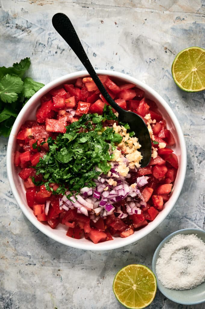 A bowl of fresh Pico de Gallo containing chopped tomatoes, red onion, garlic, and cilantro with a spoon, surrounded by lime halves, salt, and cilantro on a textured surface.