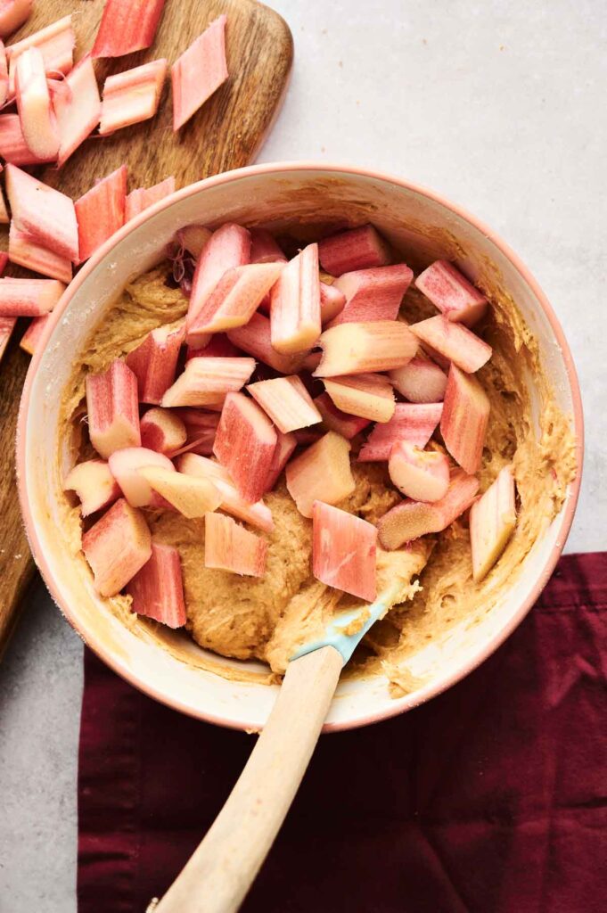 A mixing bowl with raw cake batter and diced rhubarb chunks on top, next to a cutting board with more rhubarb pieces.