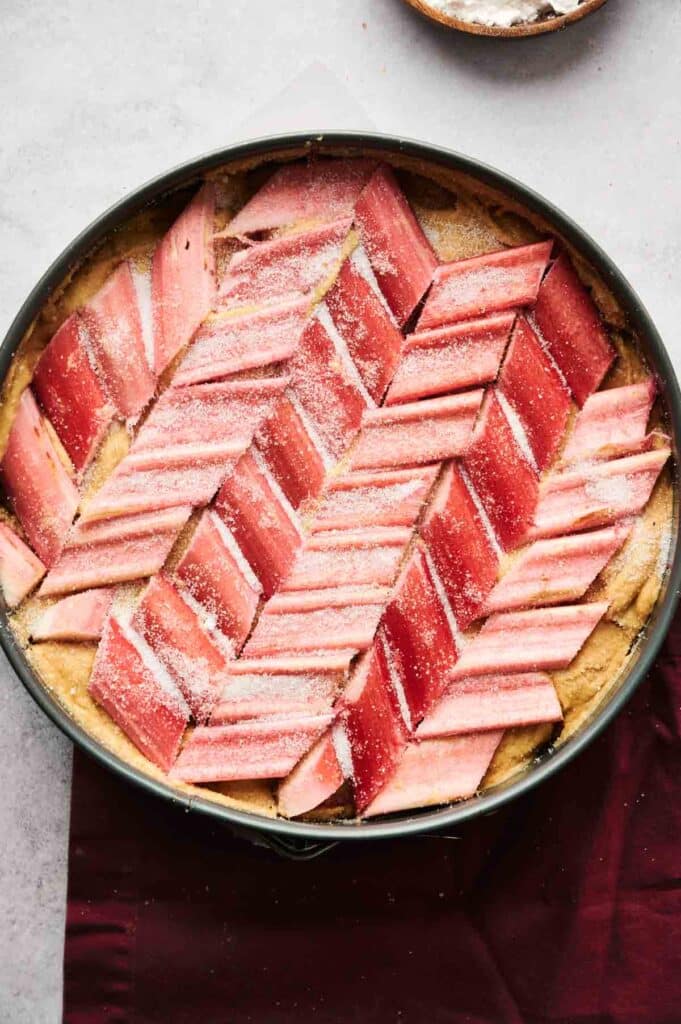 A round baking dish with uncooked rhubarb tart, arranged in a red and pink herringbone pattern, set on a light gray surface.