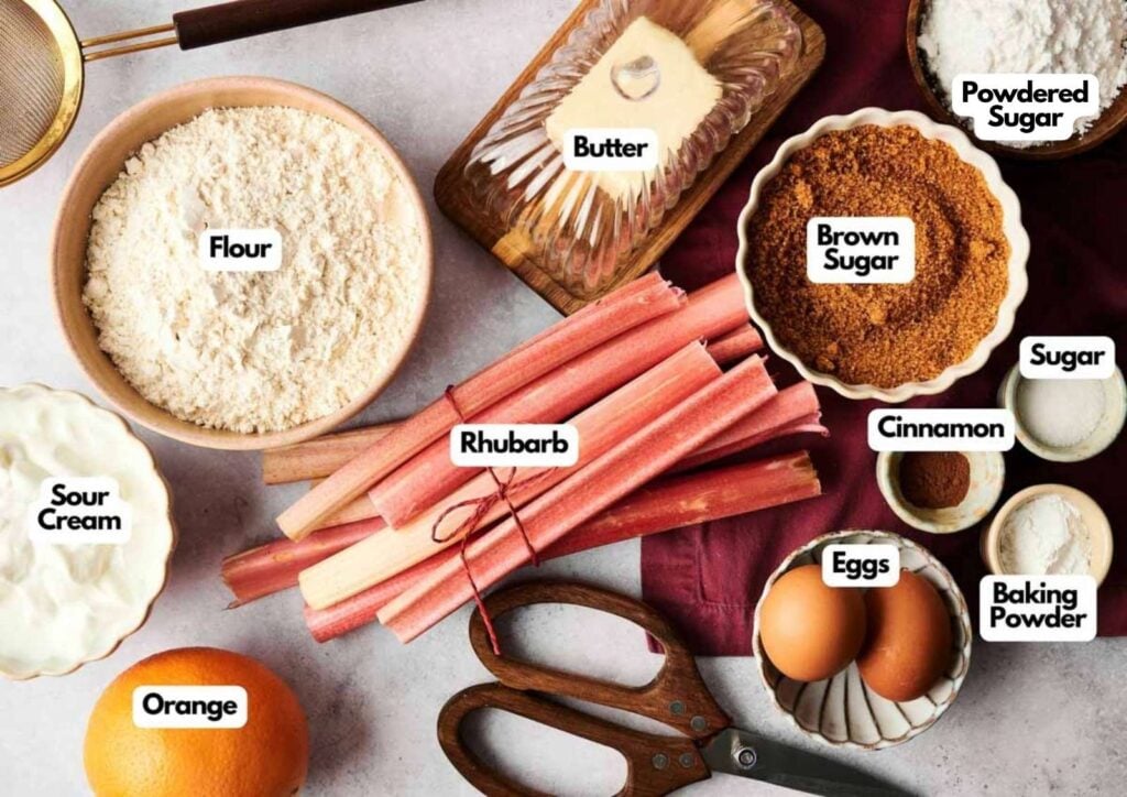 A flat lay of baking ingredients for a delicious rhubarb cake, including flour, butter, sugars, cinnamon, baking powder, eggs, sour cream, fresh rhubarb, and an orange, each labeled with white text.