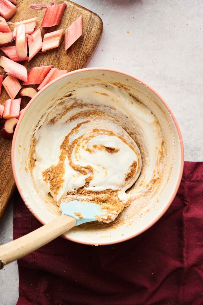 A bowl with a mixture of cream and brown batter being stirred with a blue spatula. Diced rhubarb is on a wooden board nearby.